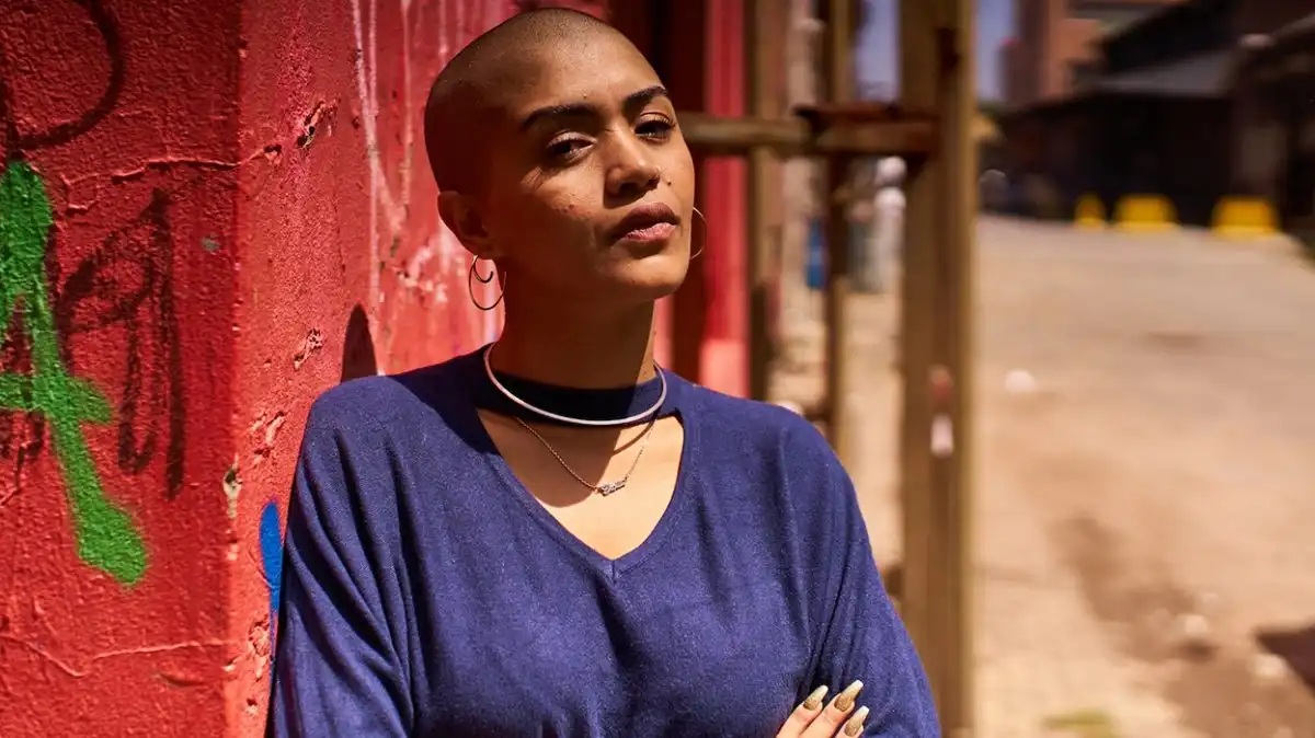 Person with a shaved head in a blue top leans against a graffiti-covered wall, with an urban street in the background.
