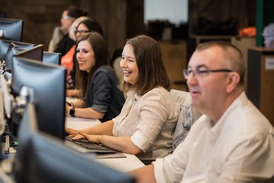 People sitting in a row at computers, smiling and engaged in a lively office environment.