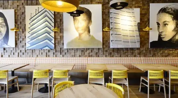 Modern restaurant interior with wooden tables, yellow chairs, and large portrait artwork on textured walls. Warm, inviting lighting.