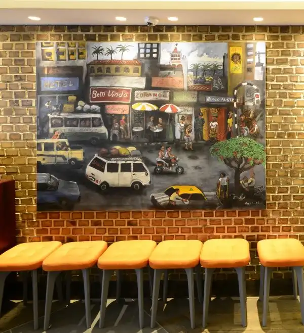 A vibrant street scene painting on a brick wall, above a row of orange-cushioned stools in a cozy setting.