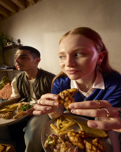 Girl in blue top holding a Nando's chicken wing, boy with Nando's on a plate in his lap