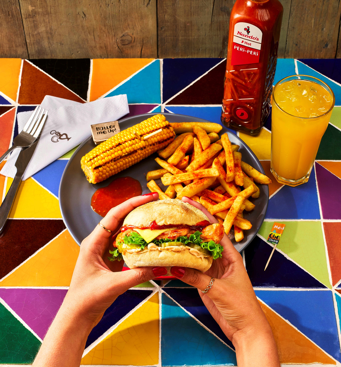 Hand holding Nando's Cheesy Chickle Burger infront of a plate of PERi chips and Corn